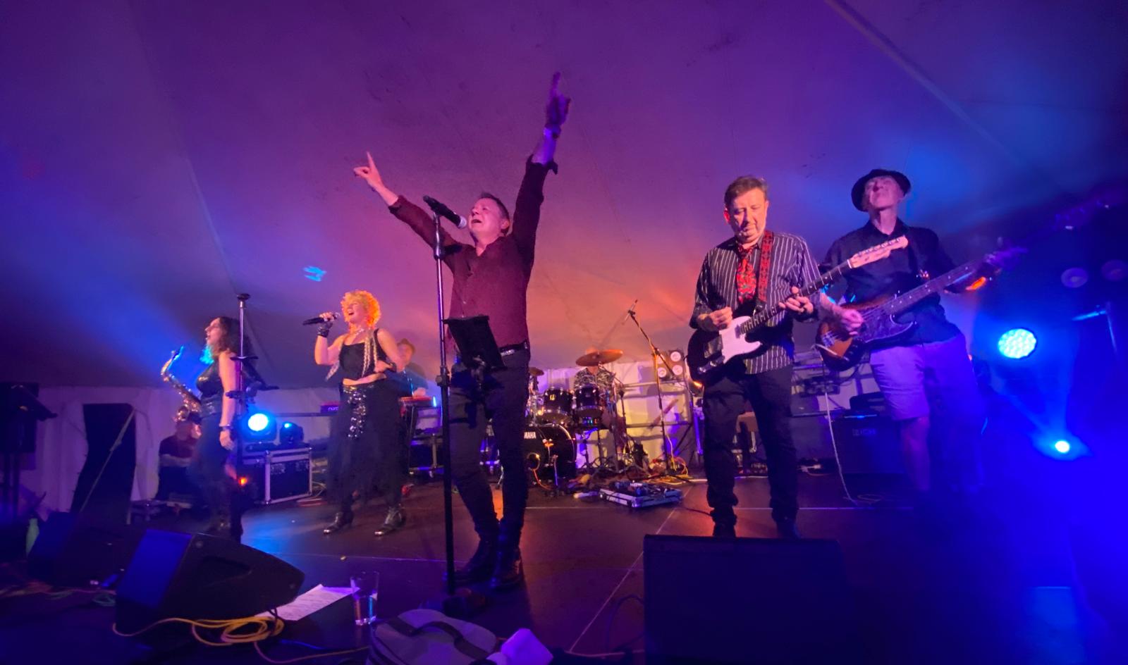 The band performing on stage with colorful lights, featuring a vocalist with a mic, a saxophonist, and musicians playing guitar and bass.